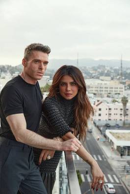 A man in a black T-shirt and a woman in a high-necked, short-sleeved black dress lean together on a balcony railing, overlooking Los Angeles.